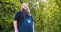 Mann mit langem Bart in blauer Latzhose und schwarzem T-Shirt vor grünen Büschen