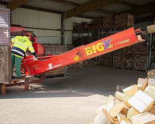 Person bedient eine rote Holzspaltungsmaschine in einer Werkstatt mit Holzstapeln