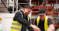 Zwei Männer in Sicherheitswesten verpacken lachend einen Karton in Lagerhalle